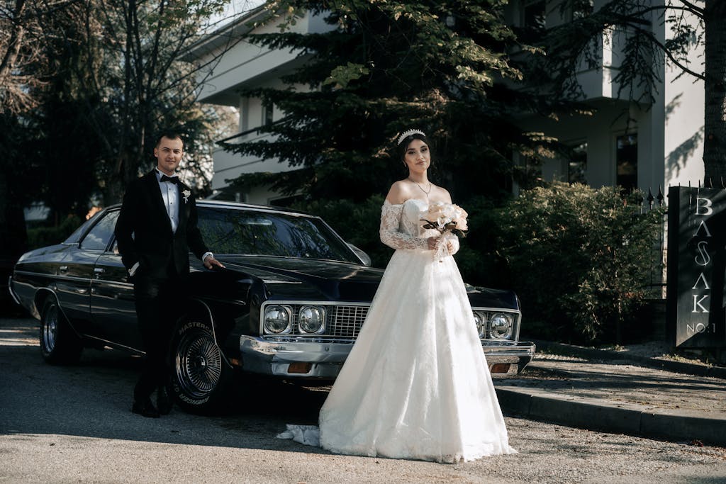Bride and groom standing by vintage car outdoors, posing elegantly on a sunny day.