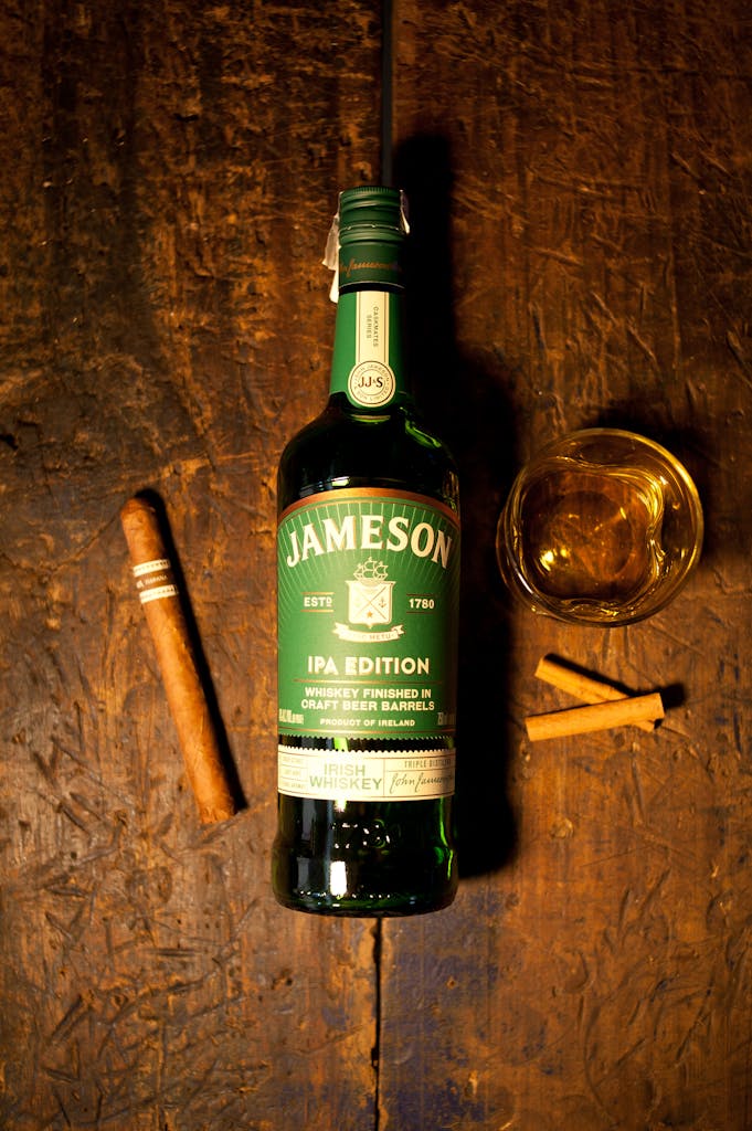 Jameson IPA Edition whiskey bottle, cigars, and glass on rustic wooden table.