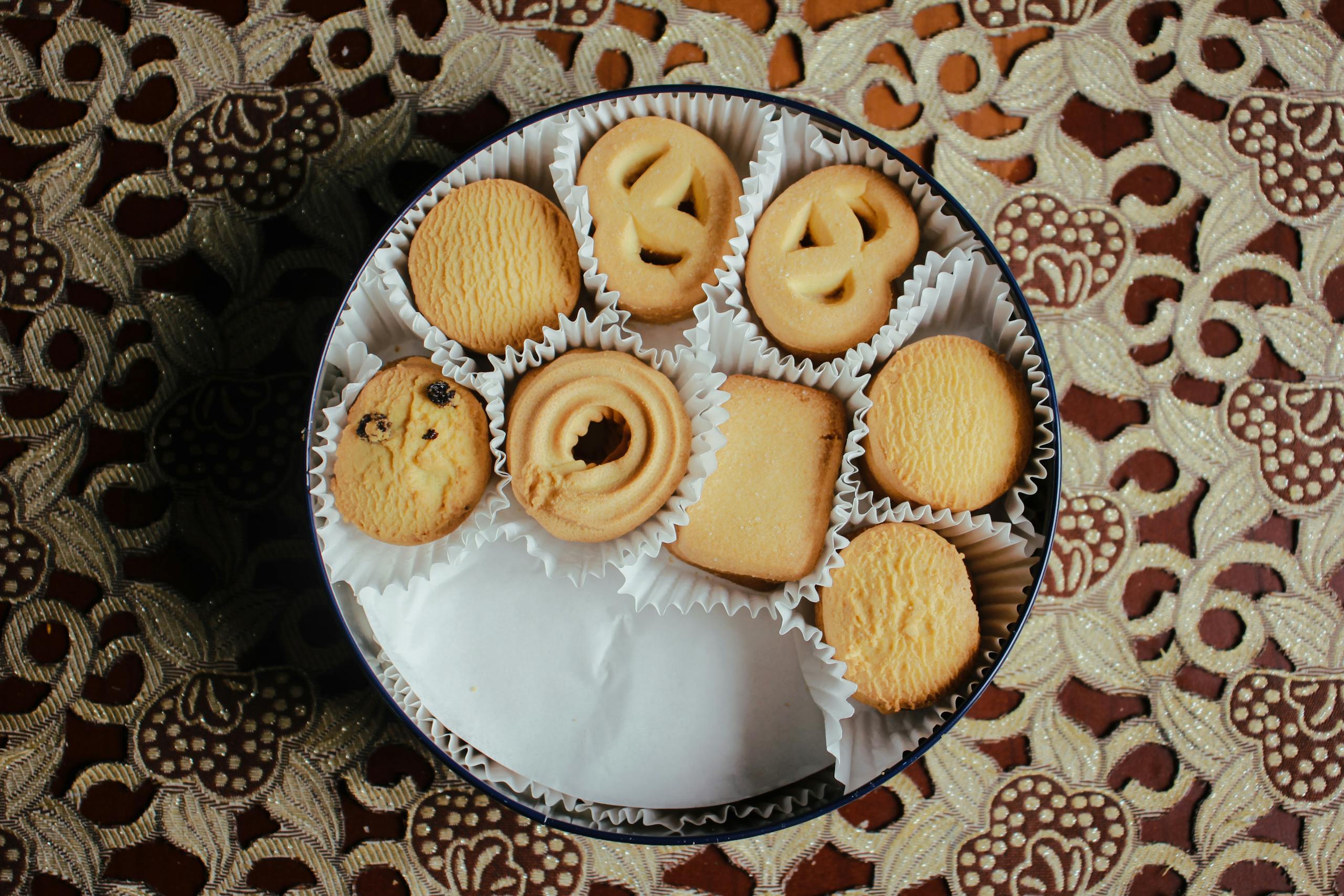 Top view of assorted butter cookies in a round tin on a decorative tablecloth.