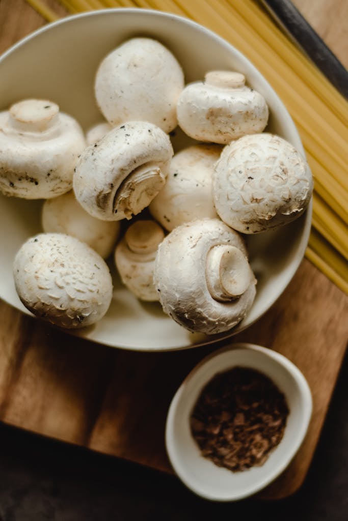Top view of fresh white mushrooms in a bowl with pasta and spices on a wooden board.
