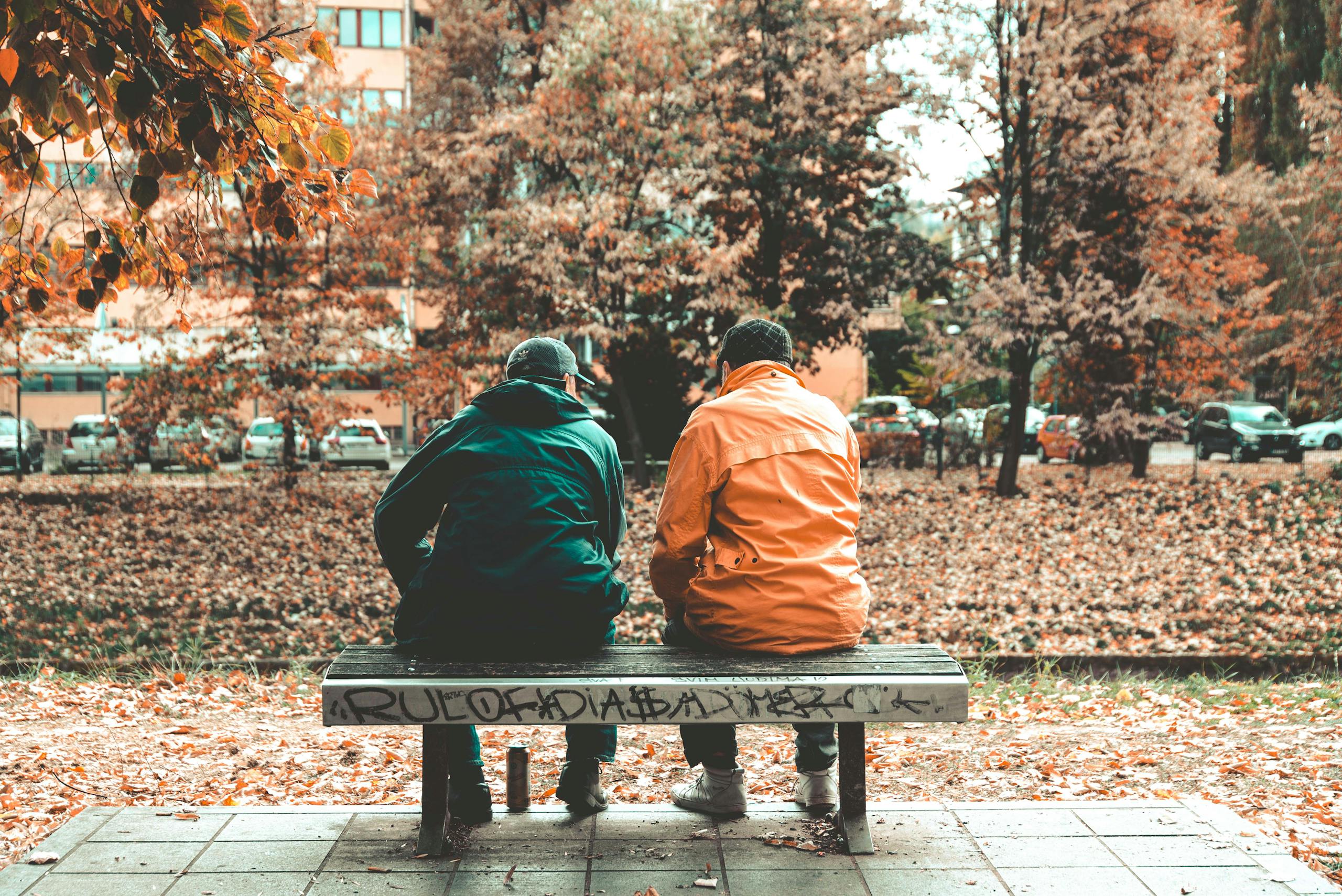 Two friends sit on a bench in an autumn Sarajevo park surrounded by fall foliage.