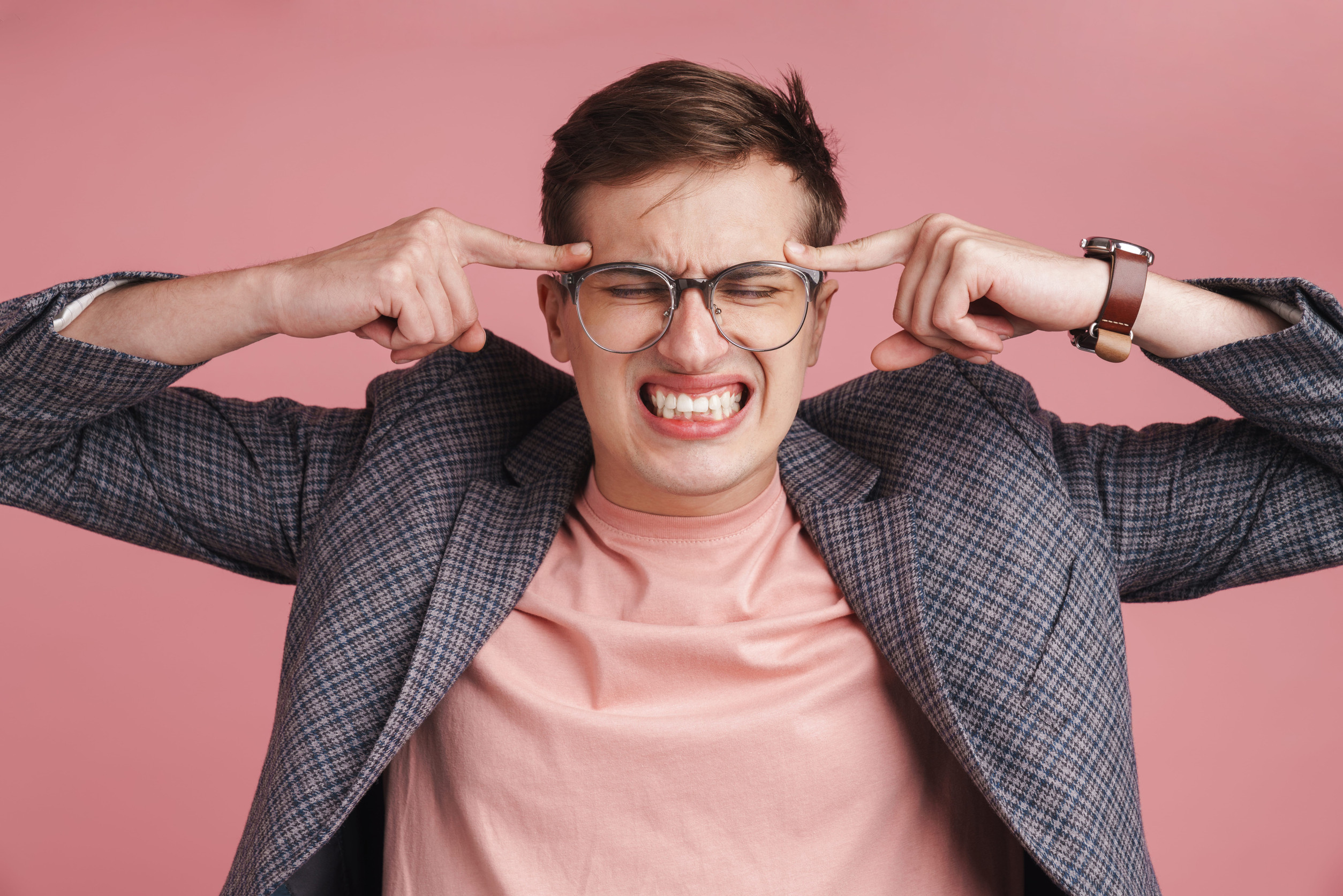 Image of a young stressed negative boy with headache in glasses isolated over pink wall background.