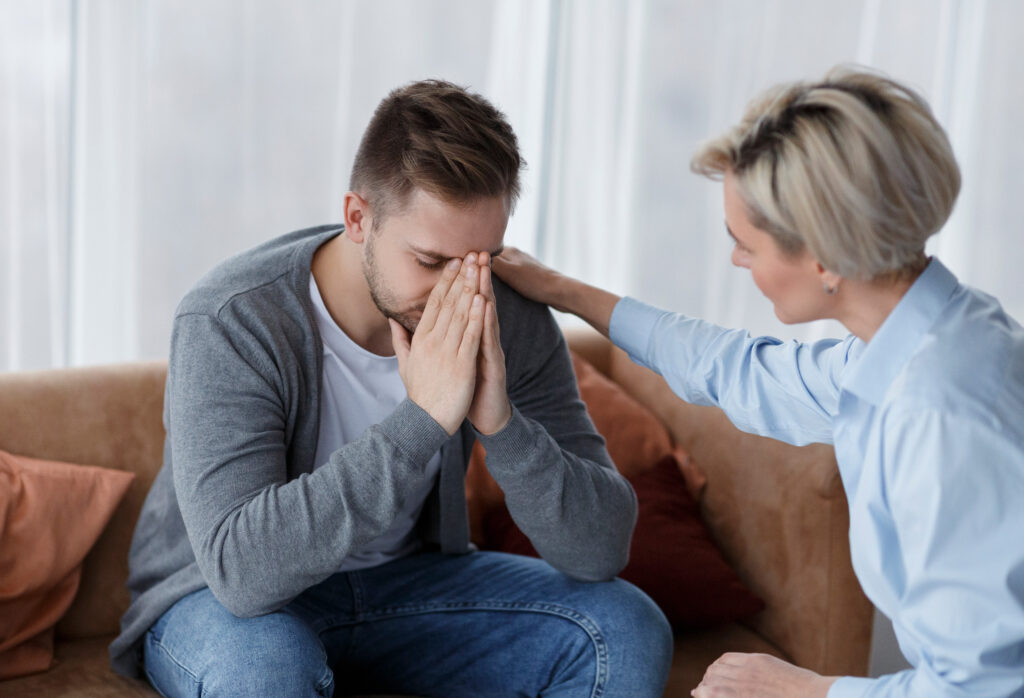 Male Depression. Professional Psychologist Comforting Crying Man During Appointment Sitting On Couch In Office. Selective Focus