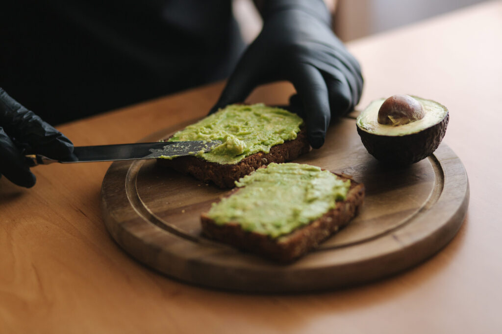 Close-up of Woman in black glowes puts guacamole or avocado spread on top of rye bread toast on wooden board at home. Vegan breakfast