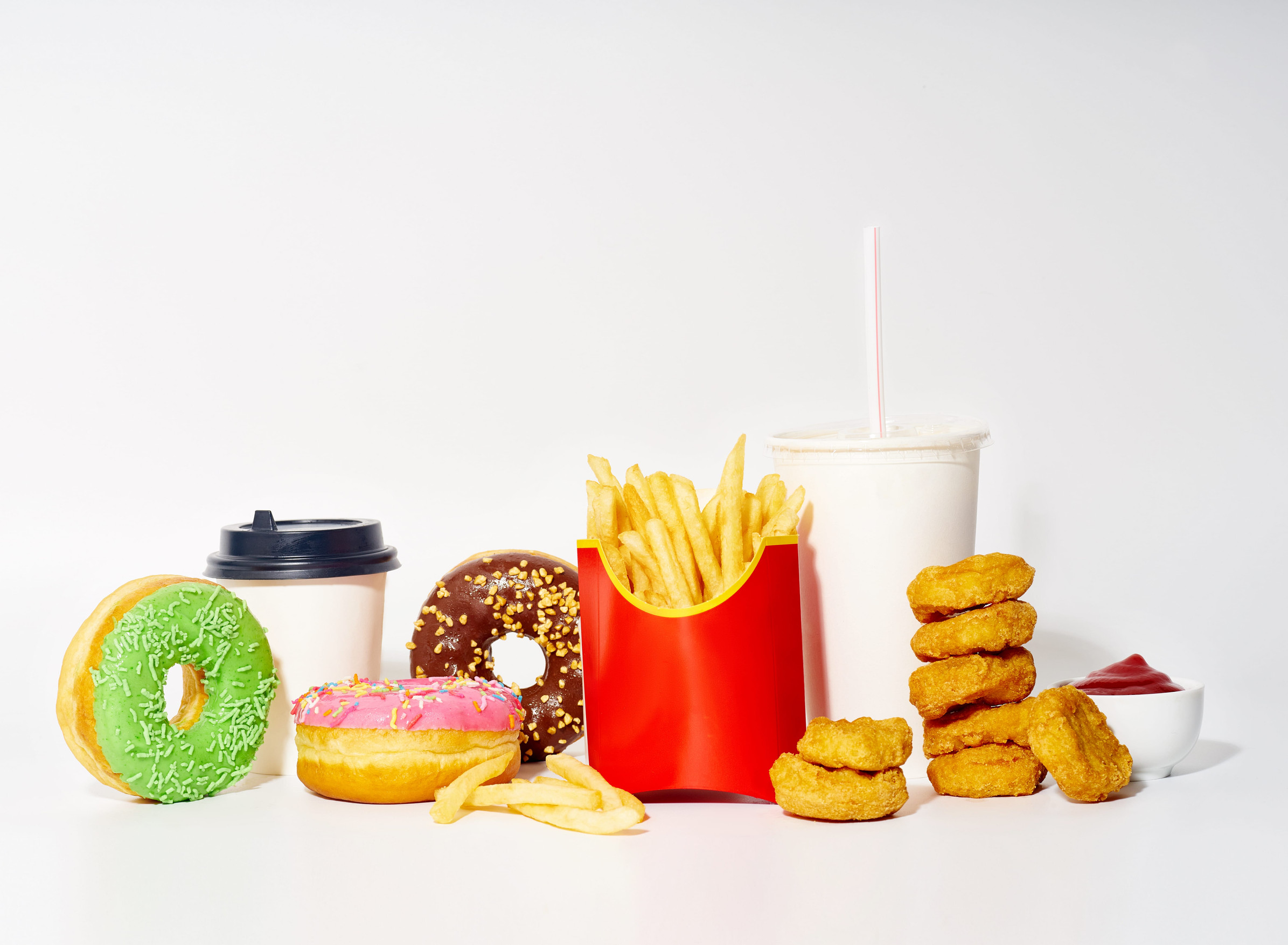 Junk food meal on white background. French fries, chicken nuggets, sweet glazed colorful donuts with drinks and sauce.