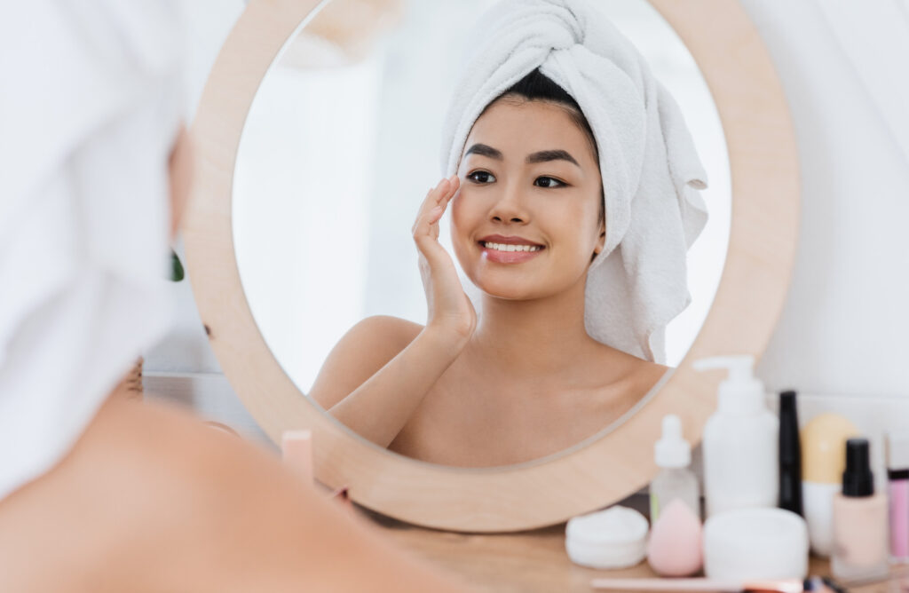 Eye care concept. Asian young woman rubbing her eye zone after shower at home, looking at mirror