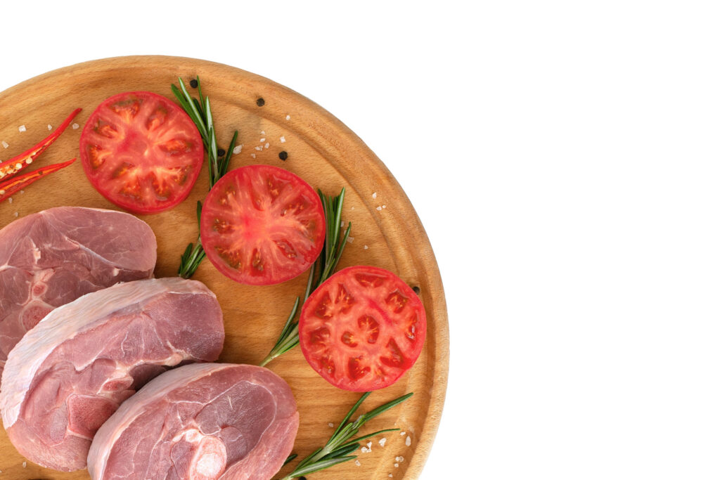 pieces of raw turkey meat, chopped leg steak, portioned barbecue pieces, on a round wooden board and and a white background with a sprig of rosemary, tomatoes and red hot pepper. isolate ...