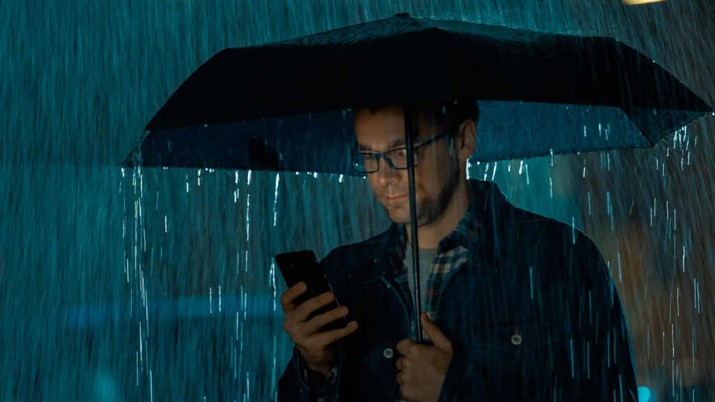 Young Caucasian Man in Glasses, Wearing a Jeans Coat and Square Shirt is Using a Smartphone Under an Umbrella. Its Dark Outside and it is Raining.