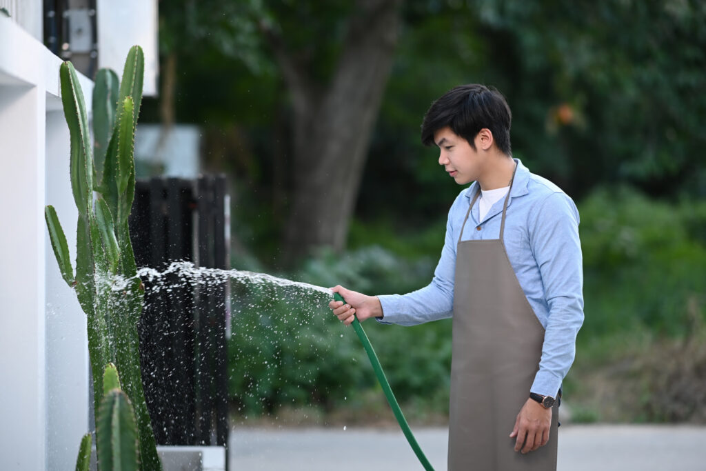A young man is holding green rubber hose and watering plants at home. Hobby lifestyle concept.