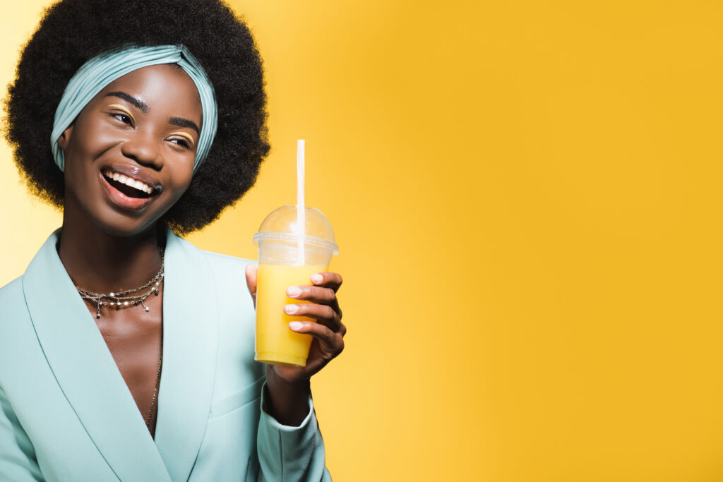 smiling african american young woman in blue stylish outfit with orange juice isolated on yellow