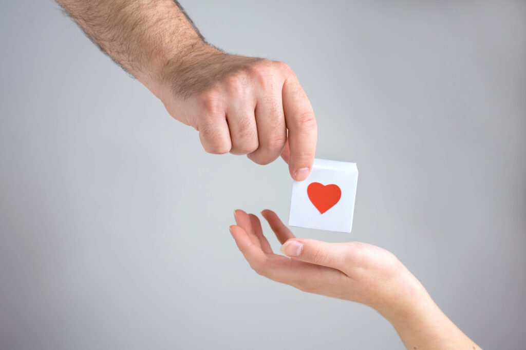 One male hand gives to one female hand a white box with a heart, simbol of love on a white background, close up.