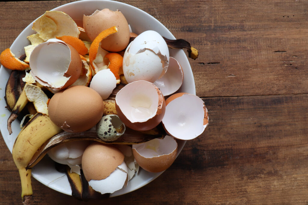 Banana skin, eggshell, and orange peels in white bowl on wooden table background. Using kitchen scraps in compost or homemade fertilizer. Zero waste concept. Top view, copy space