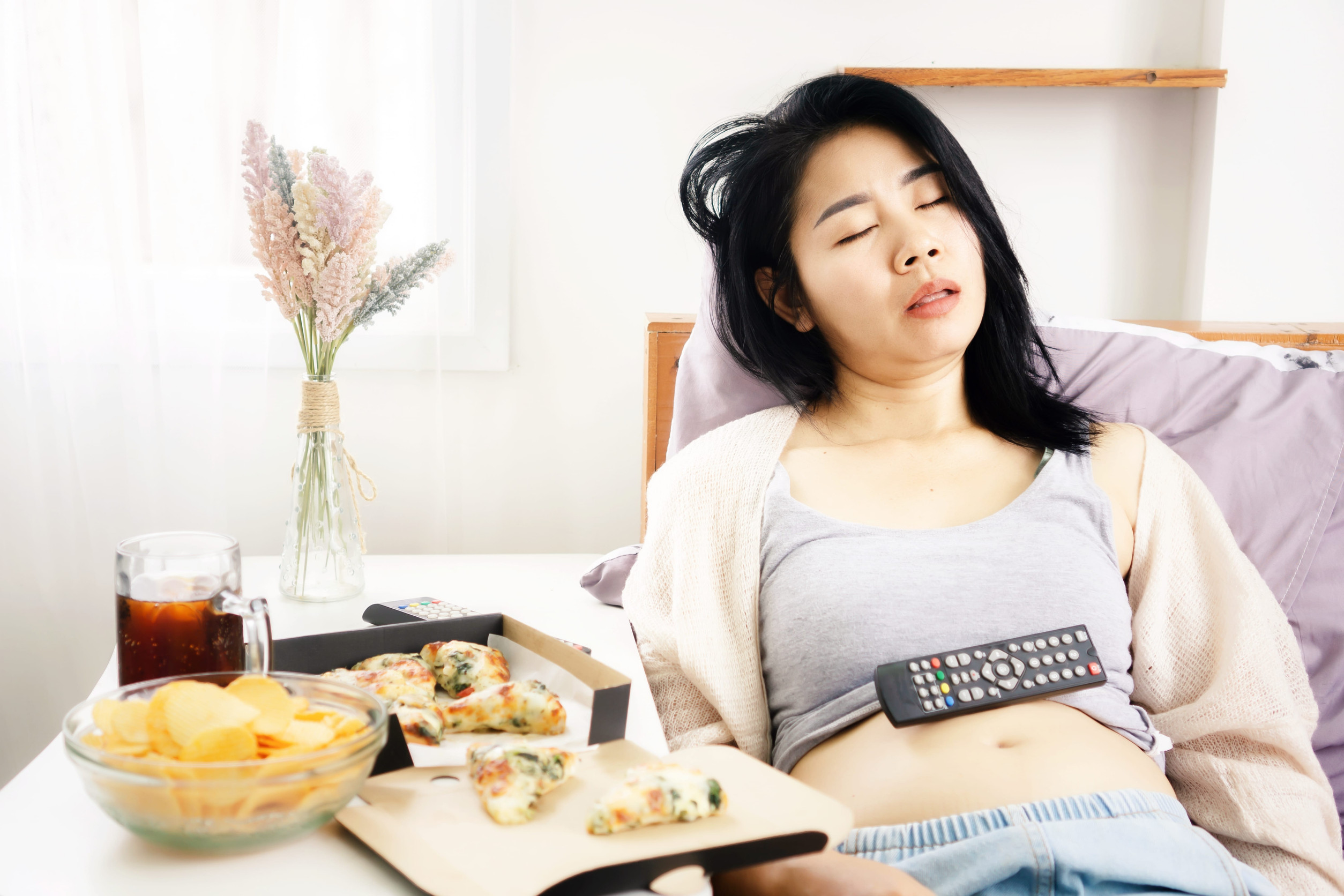 Asian woman sleeping in bed after eating pizza, potato chips and soda with Tv remote on her fat belly overeating junk food concept