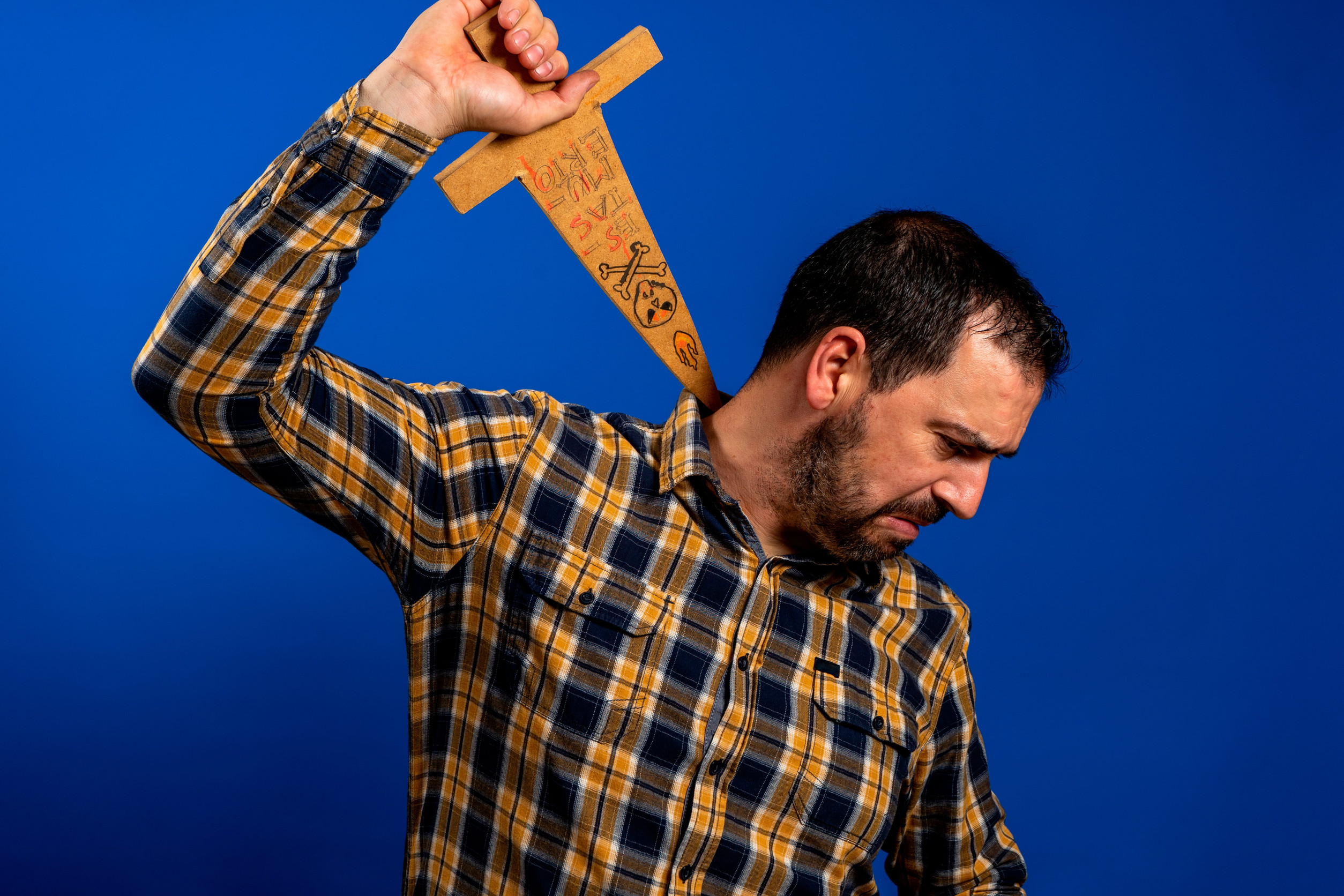 Man in a yellow plaid shirt scratching his back with a wooden sword isolated on blue studio background. Fun concept