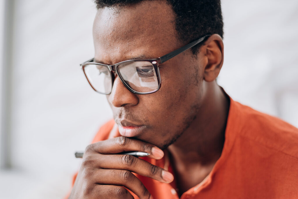 Thoughtful black man with glasses and pen in light room