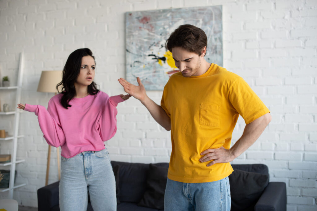 angry young woman standing with hands in air near boyfriend with refuse gesture in living room