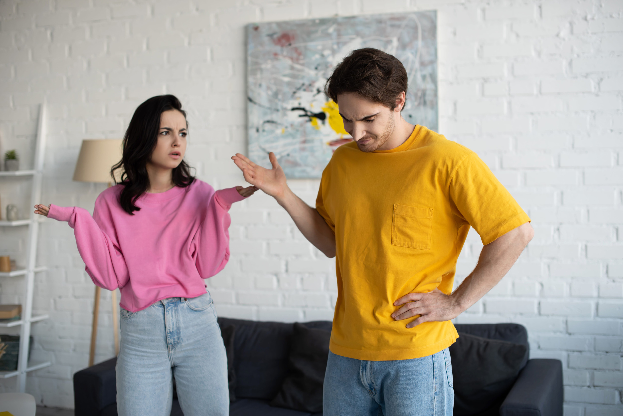 angry young woman standing with hands in air near boyfriend with refuse gesture in living room