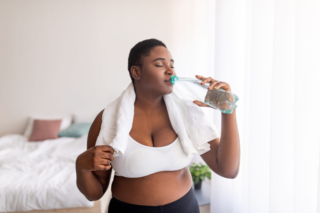 Healthy living, weight loss. Plus size black lady drinking fresh clear water from bottle after sports workout indoors