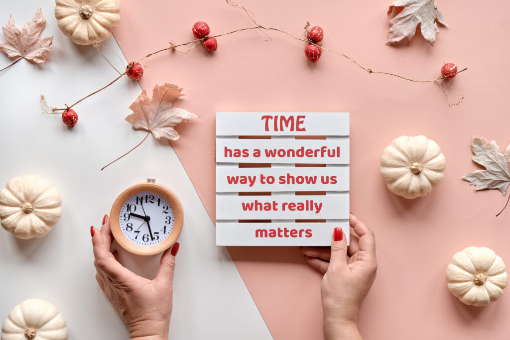 Autumntime background. Alarm clock and pallet with seasonal text in hands. Decorative pumpkins and natural garland. Layered off white and beige cream paper with dry leaves.