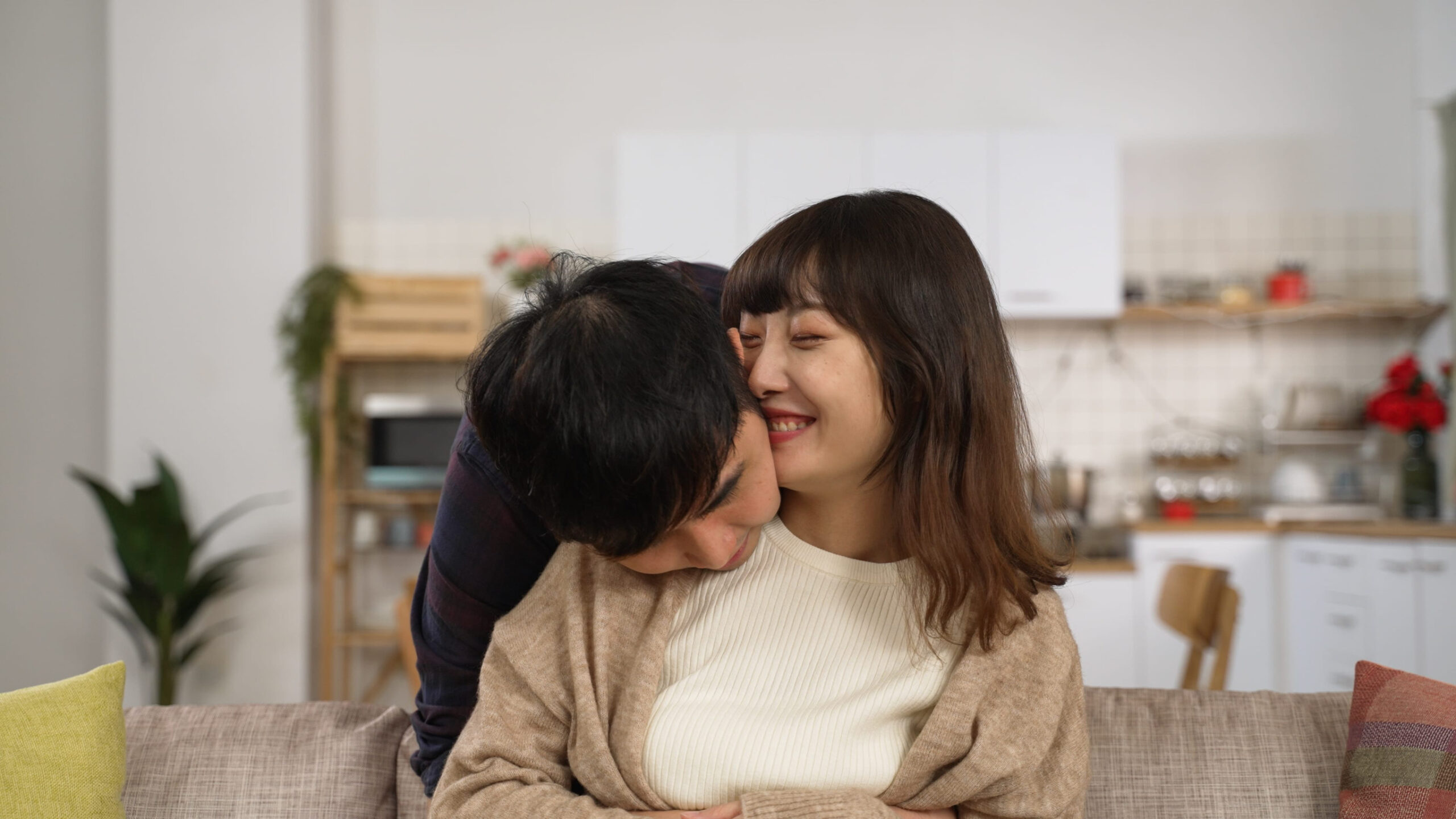 portrait of an asian husband hugging his wife from behind the couch and showing his love. they both smiling at the camera in a cozy home interior