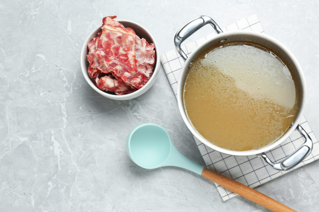 Pot with delicious broth and bowl of bones on light gray table, flat lay. Space for text