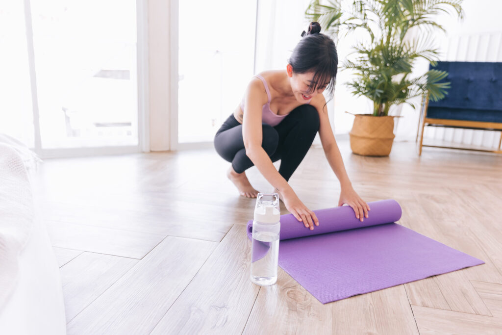 Young Asian woman wearing sportswear rolling yoga mat with water bottle, preparing for doing yoga. Healthy habits, keep fit, weight loss concepts. Focus on water bottle