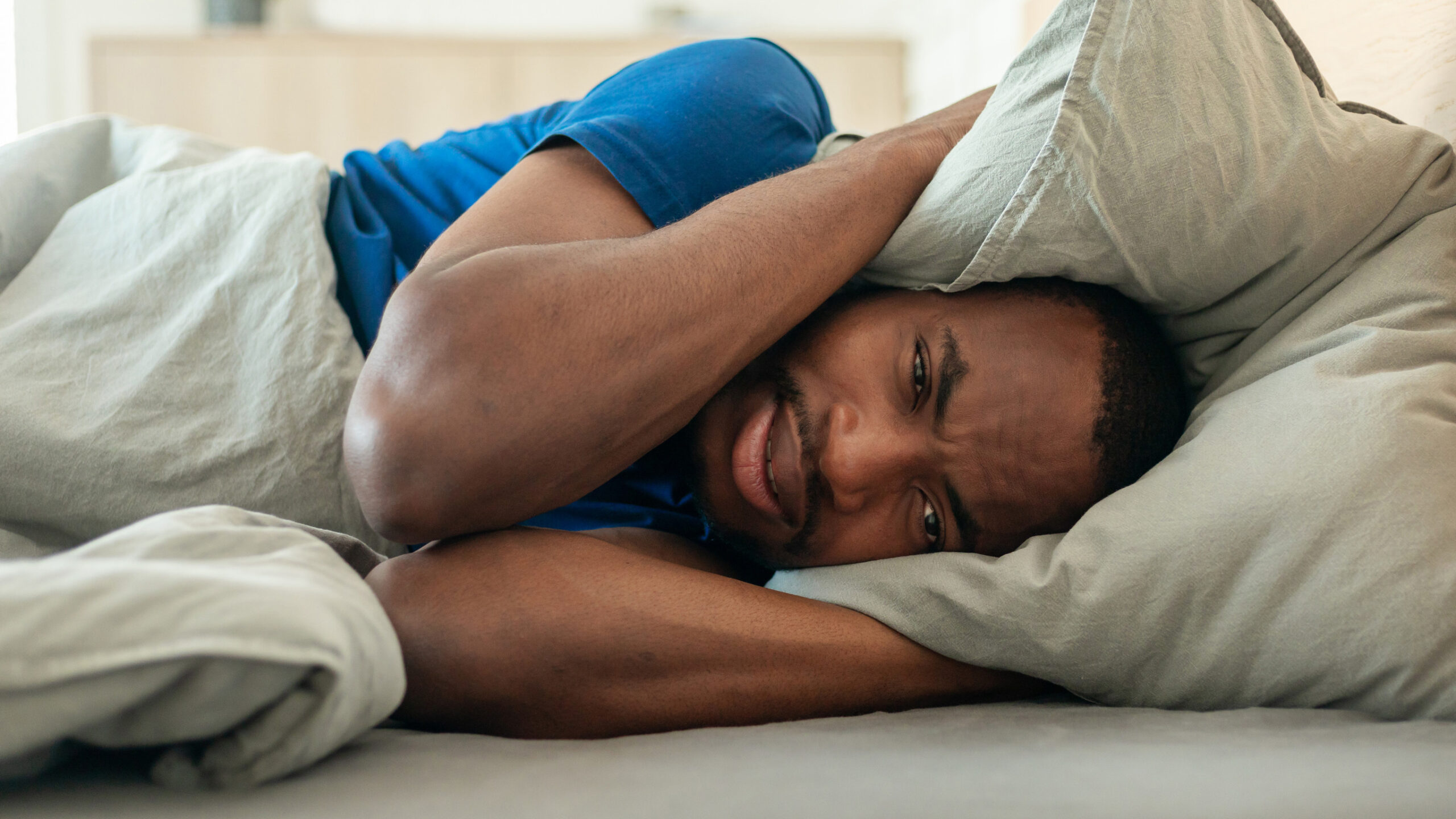 African American Guy Covering Ears With Pillow Suffering From Insomnia And Headache Looking At Camera Lying In Bed In Bedroom Indoors. Sleeping Routine And Health Concept. Panorama