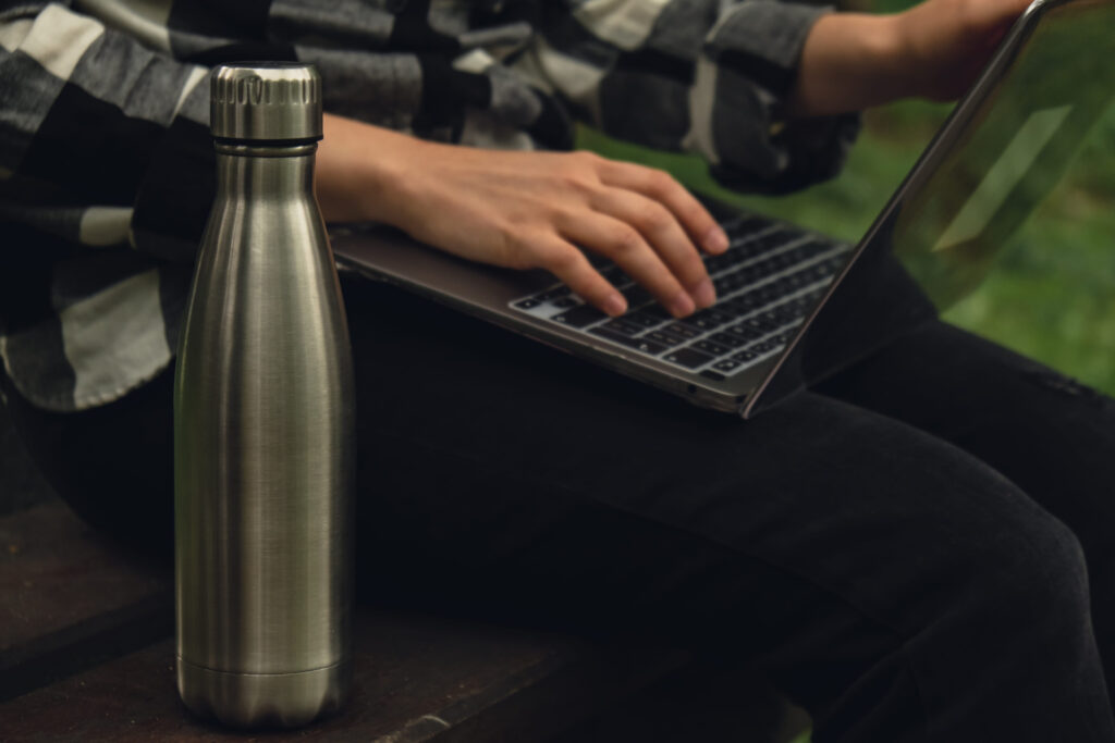 Water bottle. Reusable steel thermo water bottle on wooden bench. Sustainable lifestyle. Plastic free zero waste free living. Go green Woman student work study with laptop in park