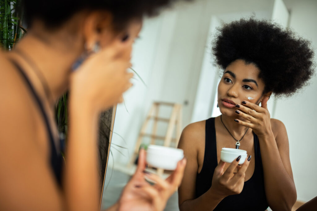 Young african american woman applying cream on face near mirror in bathroom