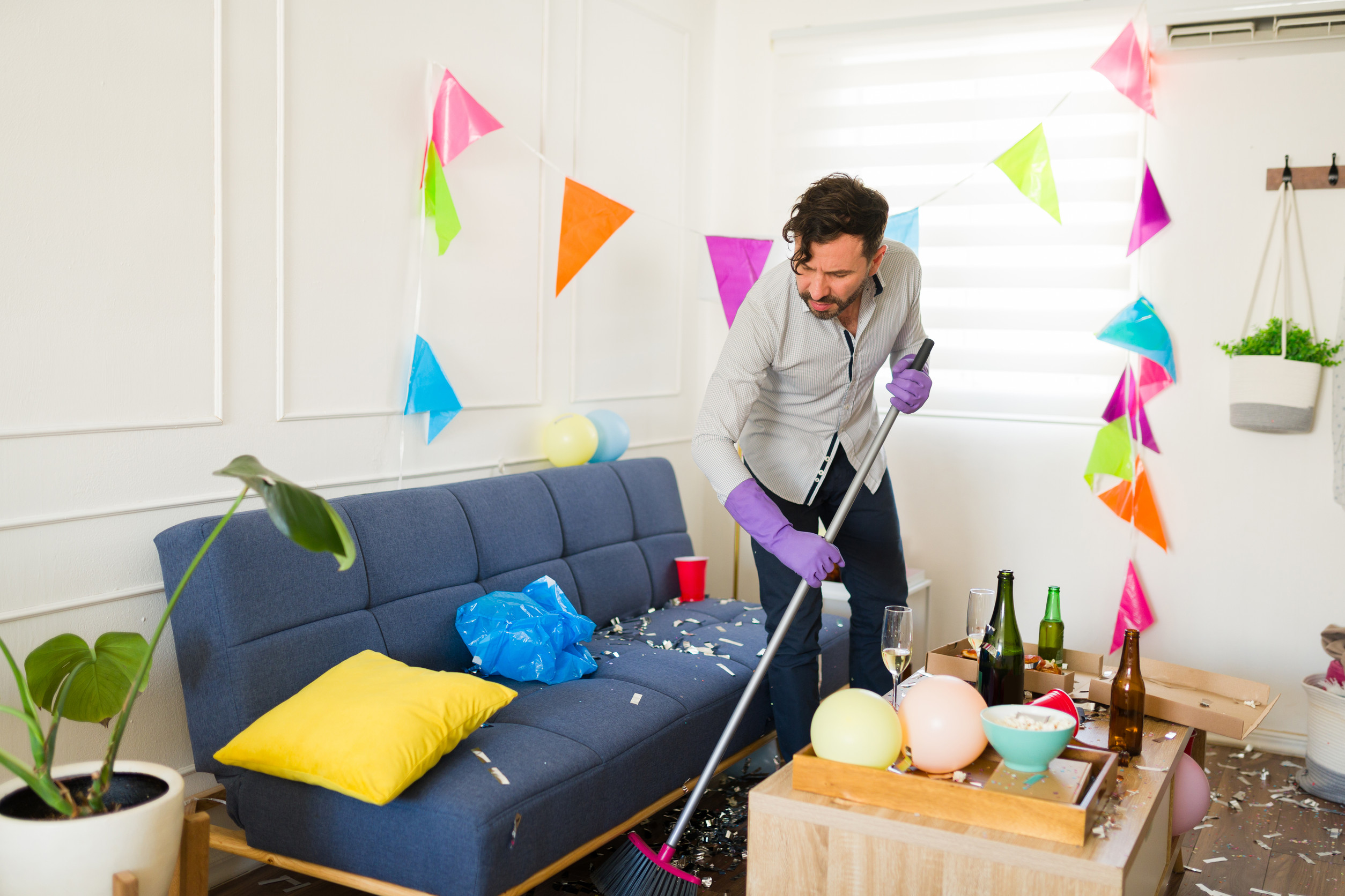 Tired man in his 30s cleaning up the trash and looking at the disaster mess at home after celebrating a party