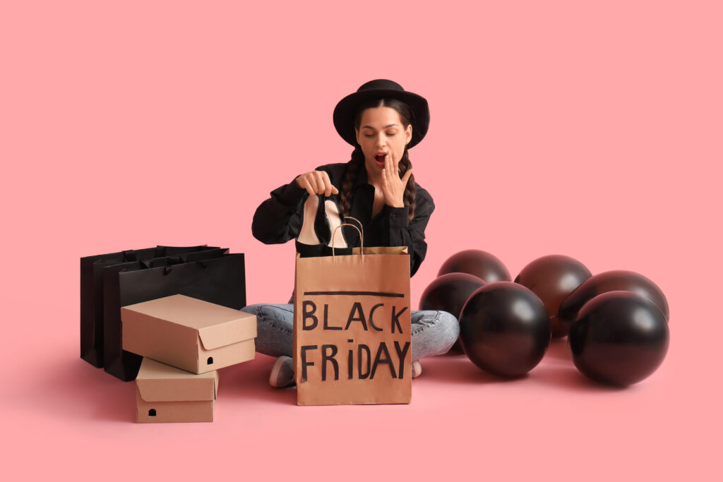 Shocked young woman with shopping bags, balloons and new heels sitting on pink background. Black Friday sale