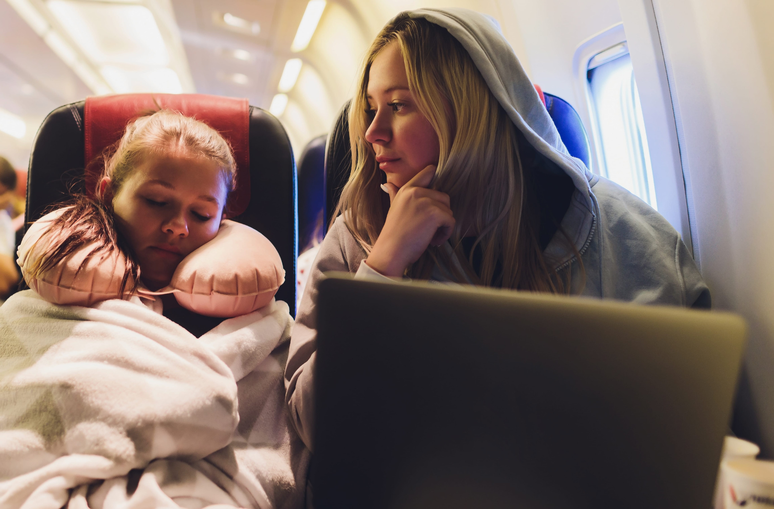 Attractive caucasian female passenger of airplane sitting in comfortable seat listening music in earphones while working at modern laptop computer with mock up area using wireless connection on board.