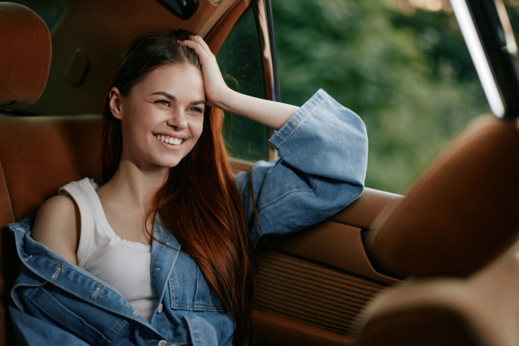 A joyful young woman with long hair relaxes in a vintage car, smiling and dressed in a denim jacket. The warm atmosphere evokes a sense of summer adventures and freedom.