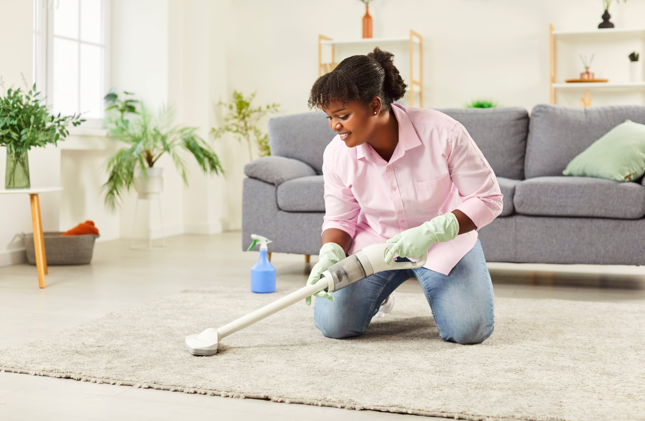 Positive African American woman, as a maid cleaning home by vacuuming the carpet on the floor. Housekeeping, hygiene, and the daily routine of maintaining a clean house living space.