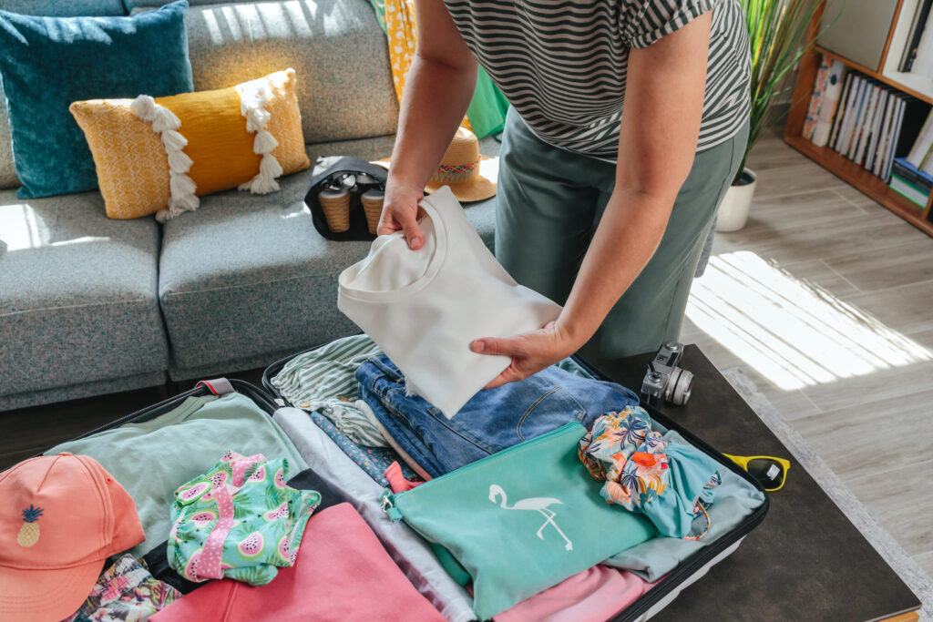 Unrecognizable woman placing clothes into suitcase with care, preparing for a summer vacation, surrounded by accessories and clothing items, creating stress free and efficient packing experience.