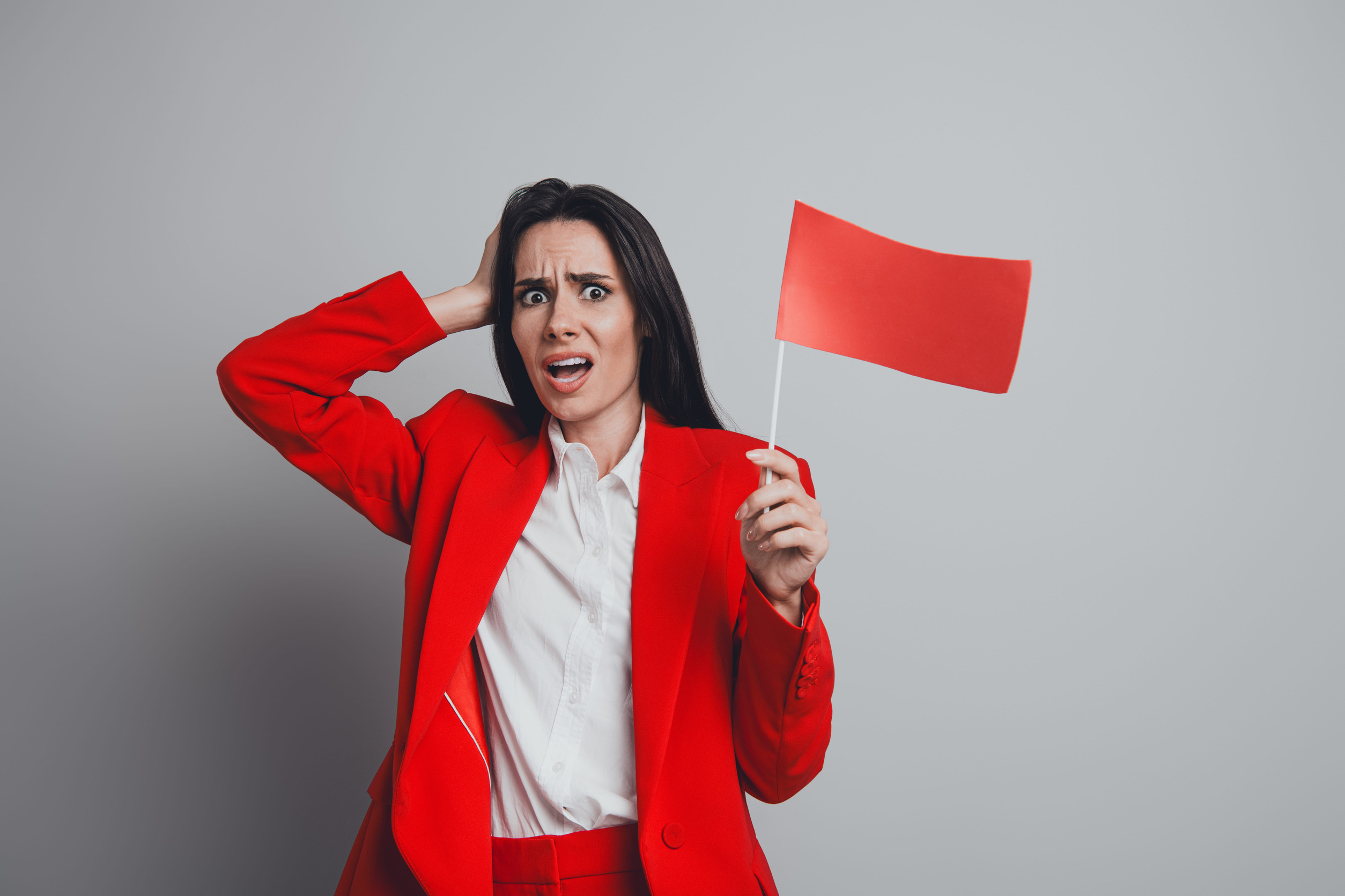 Confident businesswoman in red attire holding a red flag with a concerned expression against a neutral gray background