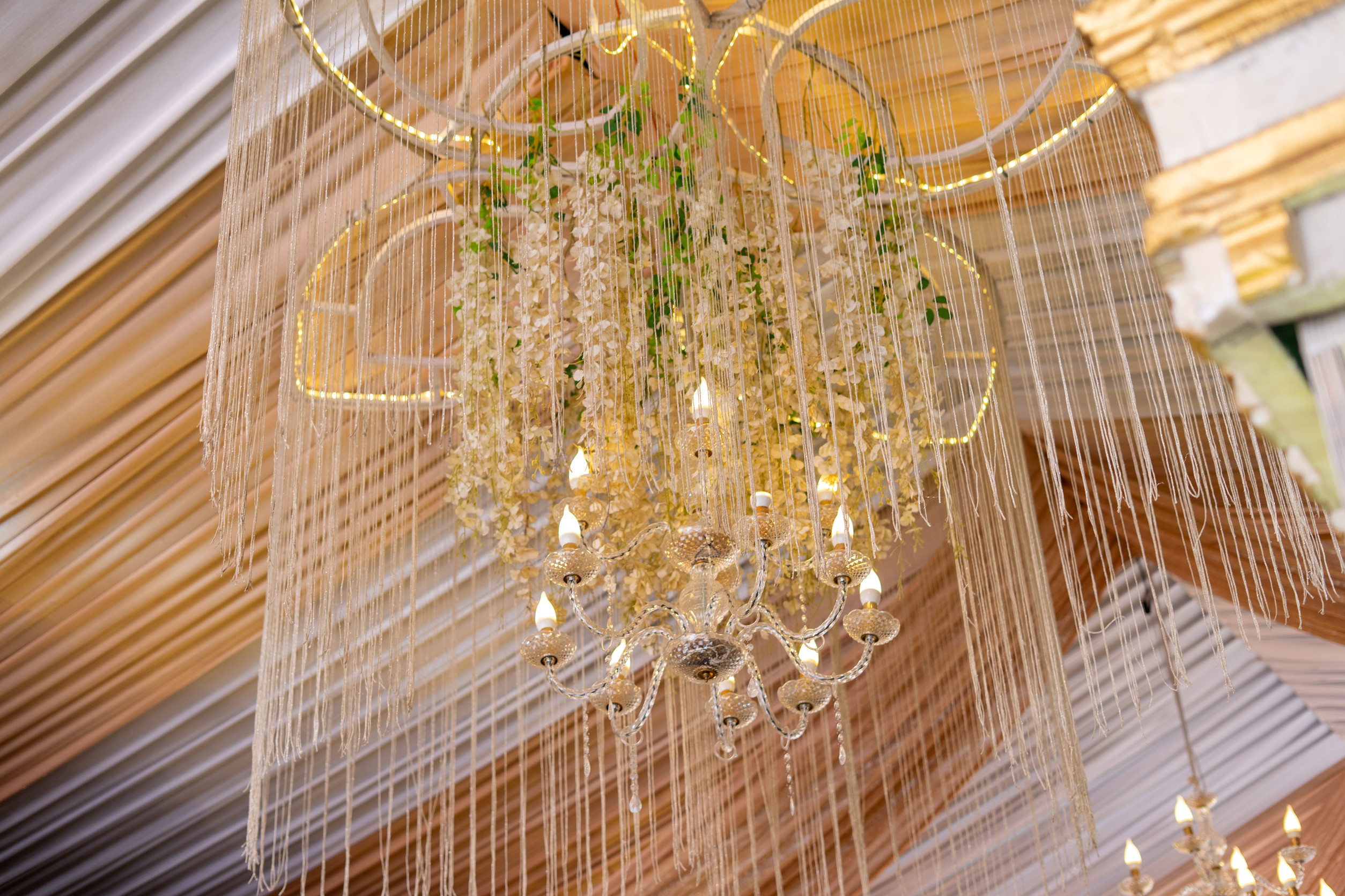 An elegant crystal chandelier adorned with delicate white hanging flowers and shimmering decorative strings, set against a background of softly draped golden and white fabric.