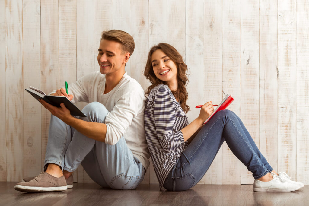 Beautiful young couple writing in a notebook sitting on wooden floor at home