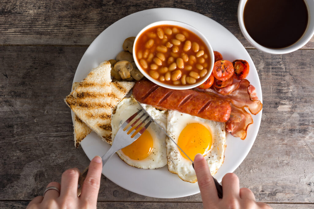 Woman eating a traditional english breakfast with fried eggs, sausages, beans, mushrooms, grilled tomatoes, bacon, coffee and orange juice, on wooden table