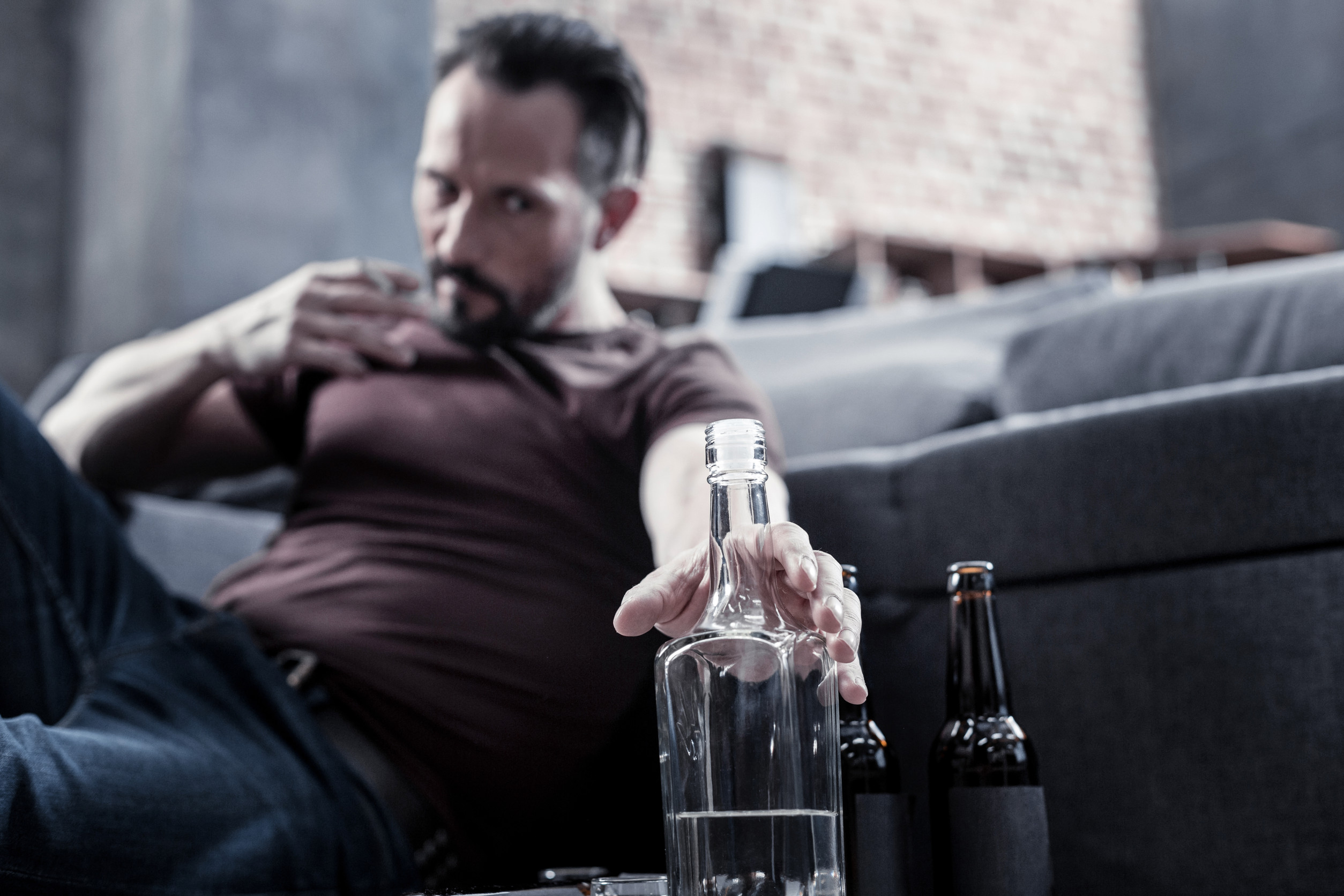 I need it. Unhappy cheerless brutal man sitting on the floor and reaching for the bottle while being addicted to alcohol