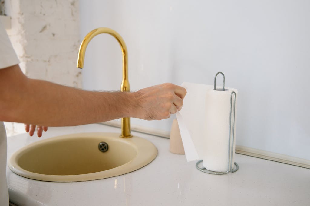 A bathroom sink with a golden faucet and hand reaching for a paper towel in a clean setting.