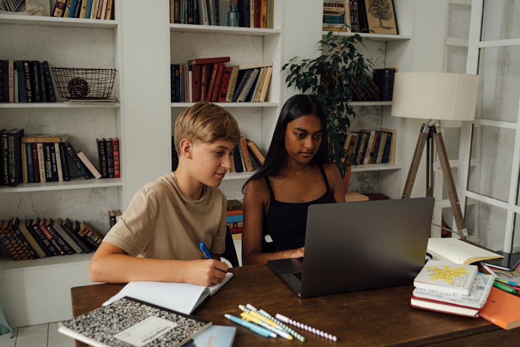 A boy and girl study together using a laptop in a home library setting.
