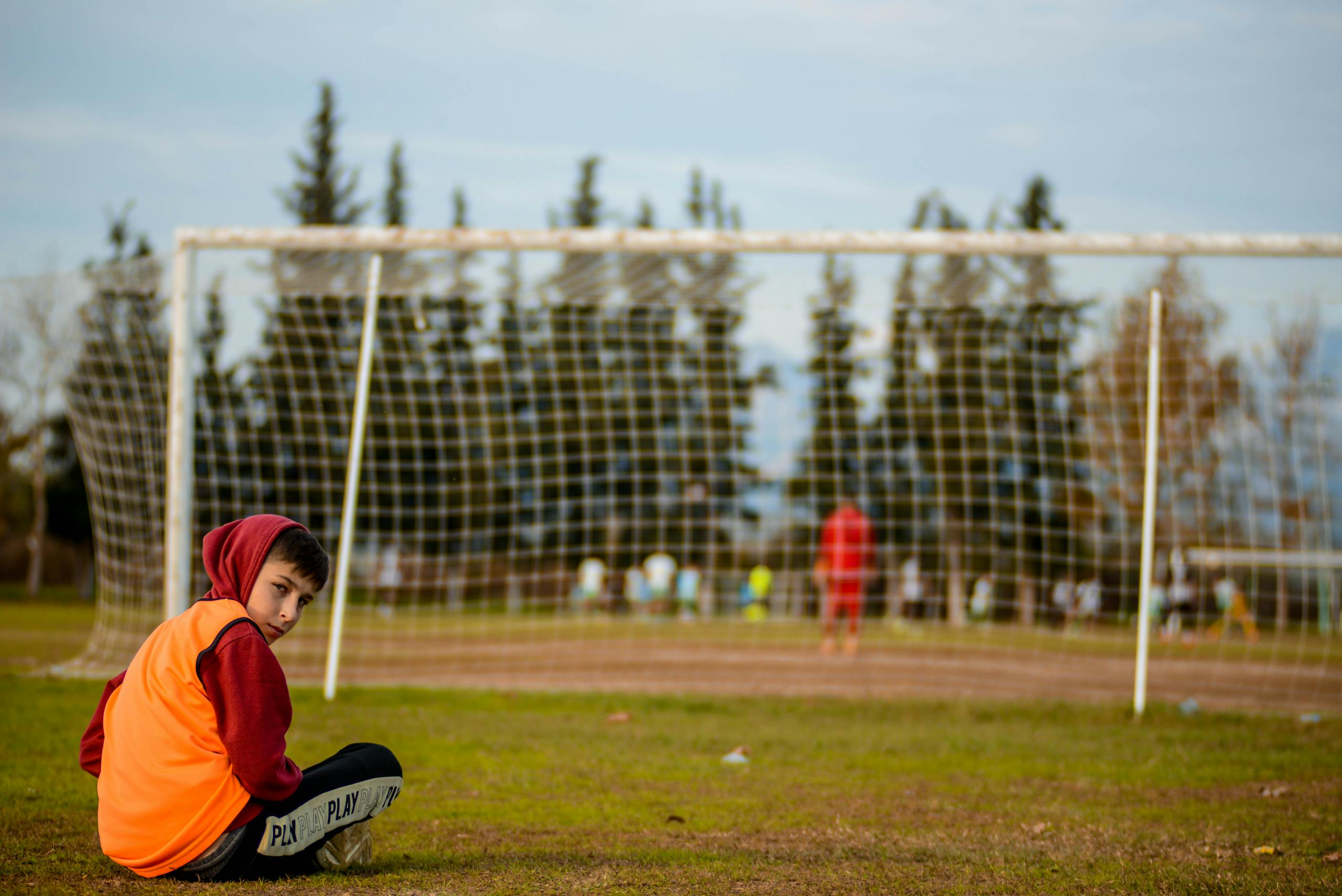 A boy sits cross-legged on a football field, wearing a hoodie and an orange vest.