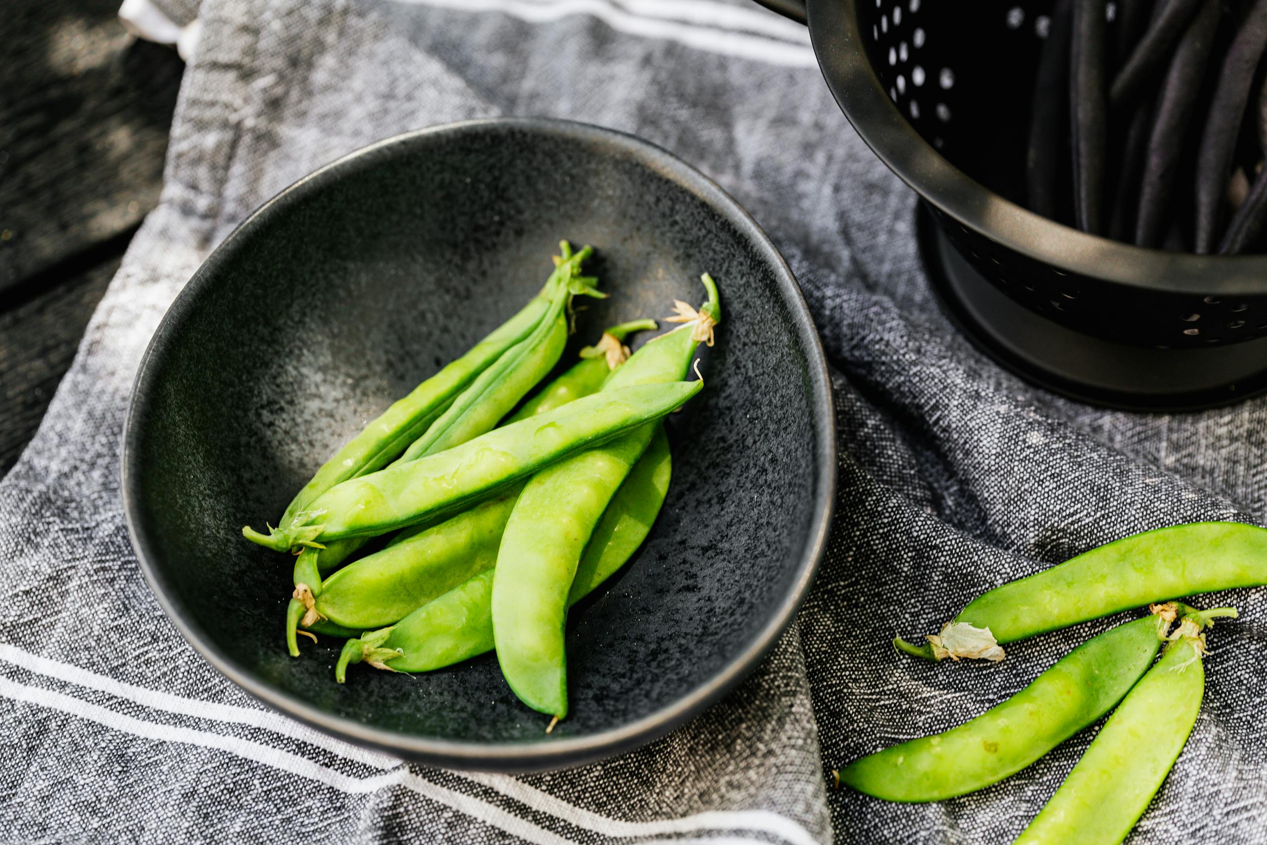 A close-up of fresh green snap peas in a black bowl on a rustic fabric background.