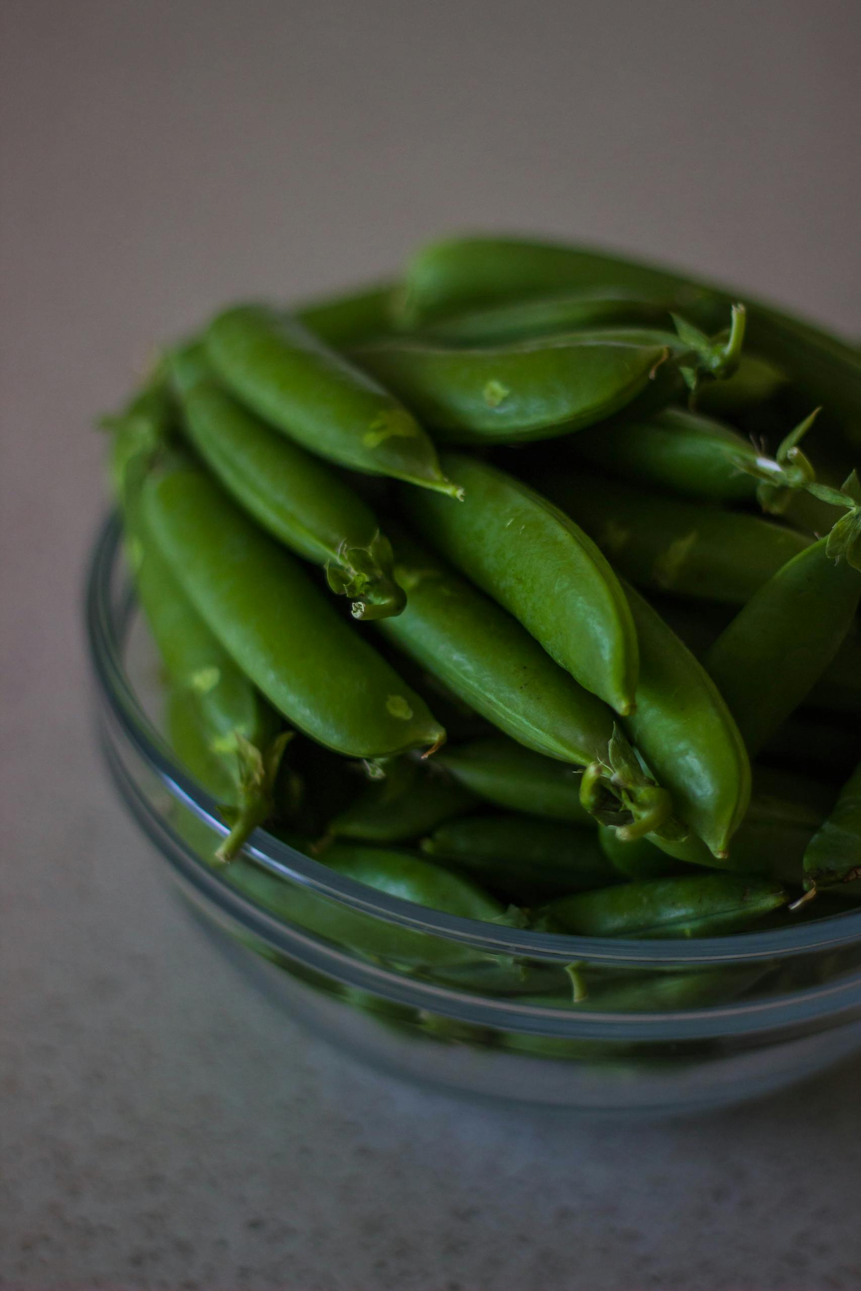 A close-up of fresh green snap peas in a glass bowl on a table.