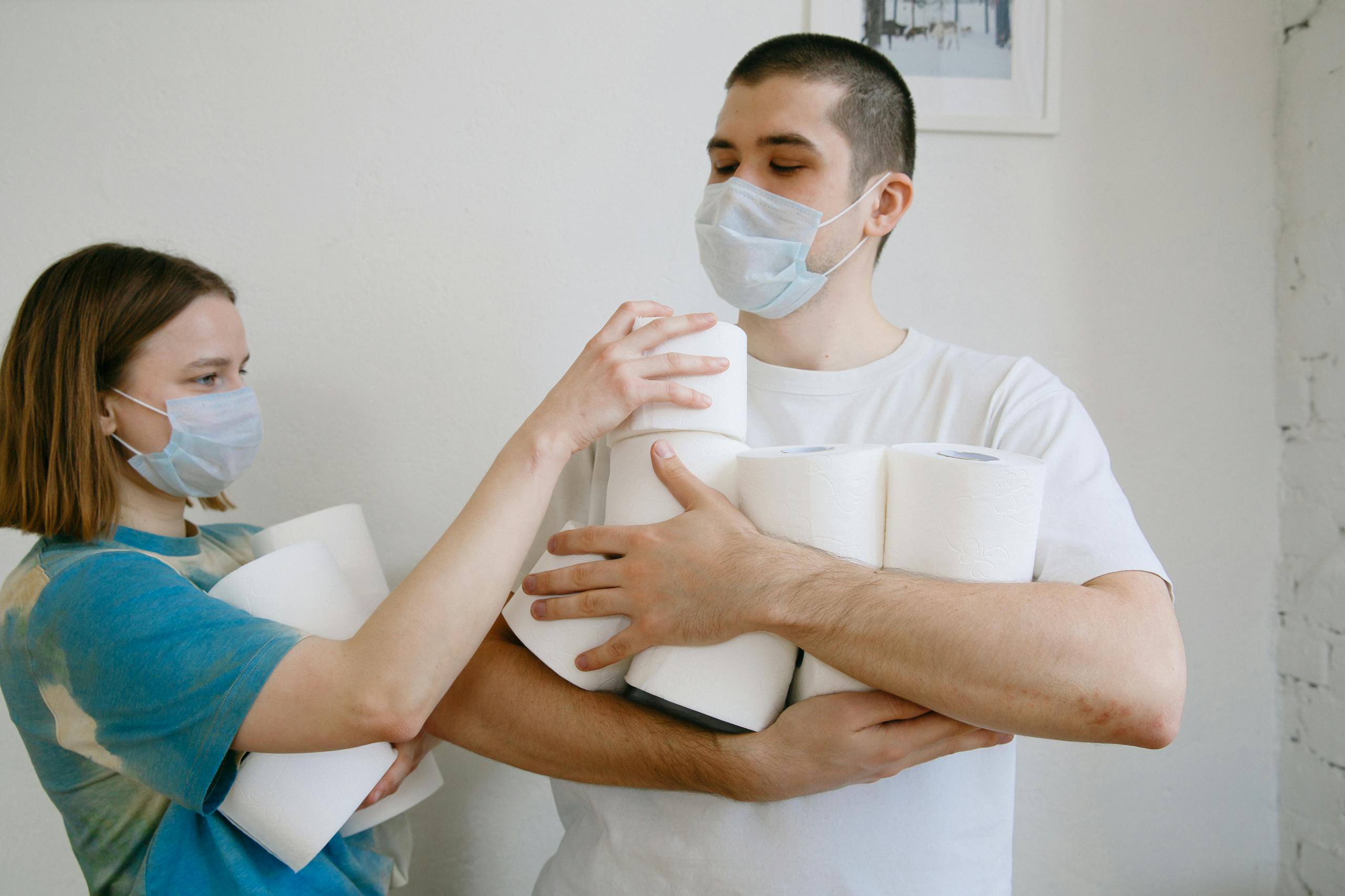 A couple at home wearing face masks, holding toilet paper rolls during quarantine.