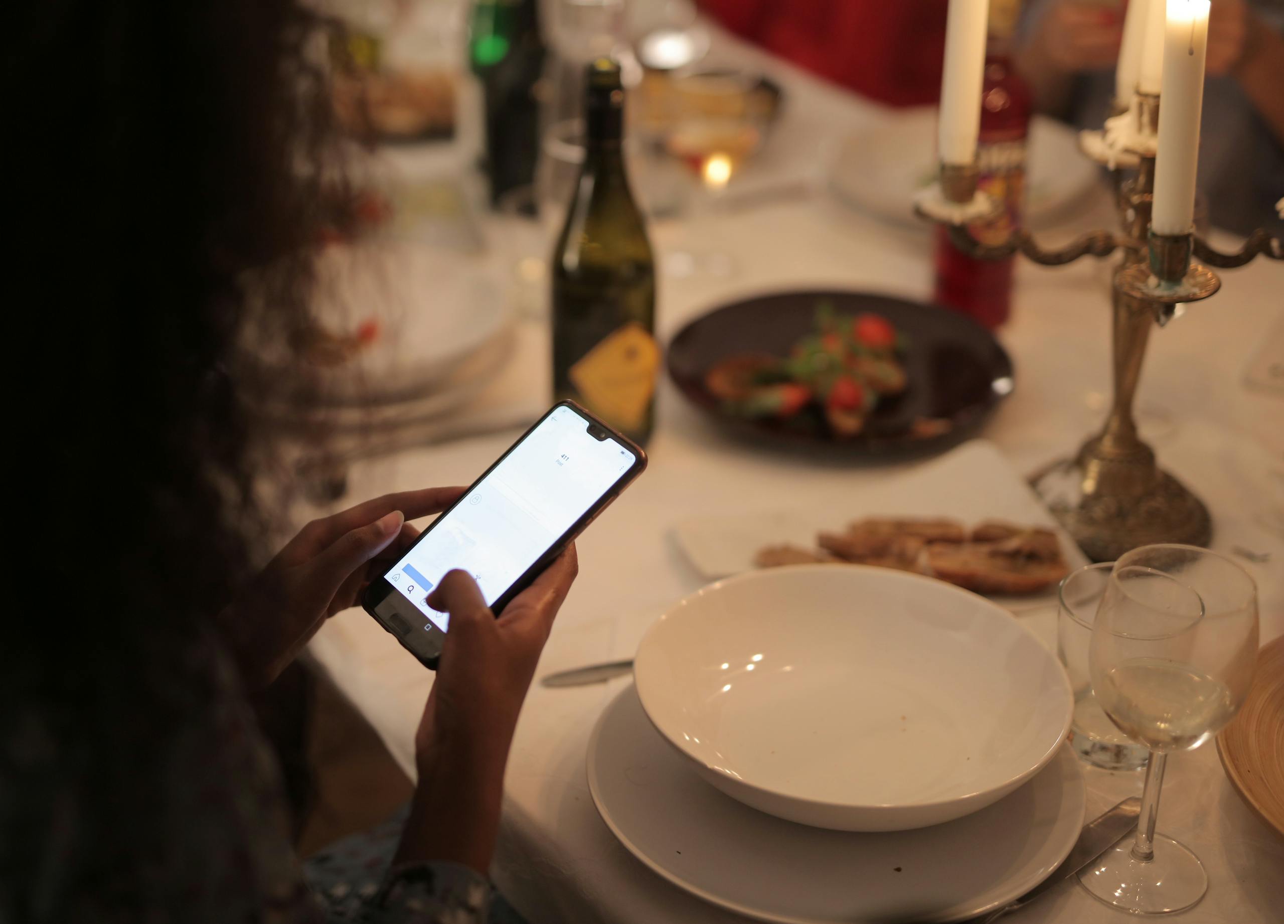 A cozy dinner setting with a woman using a smartphone at a candlelit table.