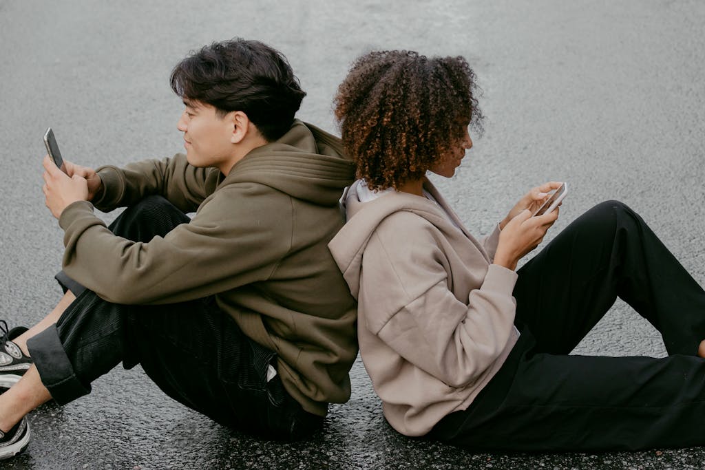 A diverse young couple sits back to back using their smartphones, depicting modern communication.