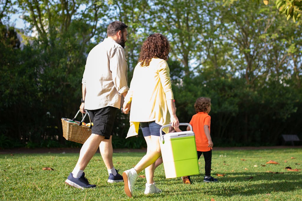 A family with a child carrying picnic essentials walking in a sunny park.
