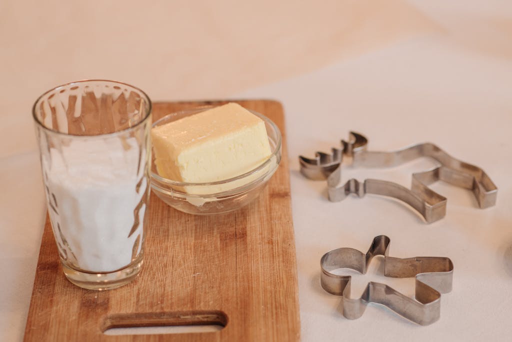 A glass of milk and butter bowl placed on a wooden board with cookie cutters, ready for baking.